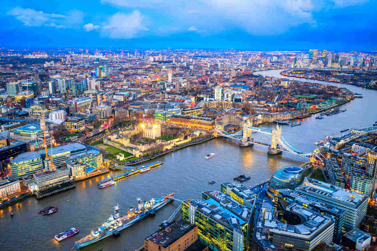 A panoramic aerial view of London at twilight, showcasing the River Thames winding through the cityscape with the Tower Bridge and the HMS Belfast