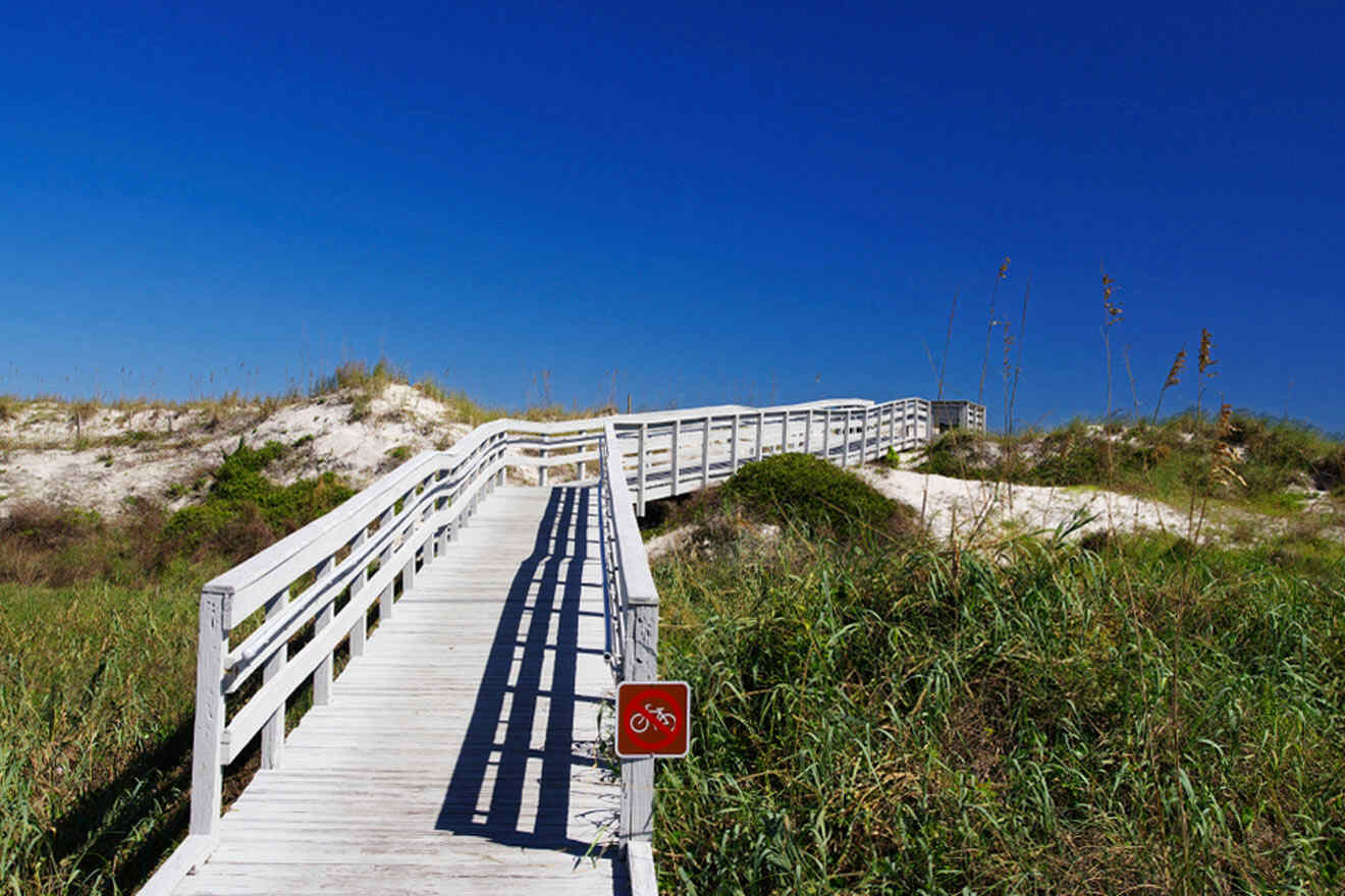 A wooden boardwalk leads through grassy dunes under a clear blue sky. A sign with a bicycle icon indicates no cycling is allowed.