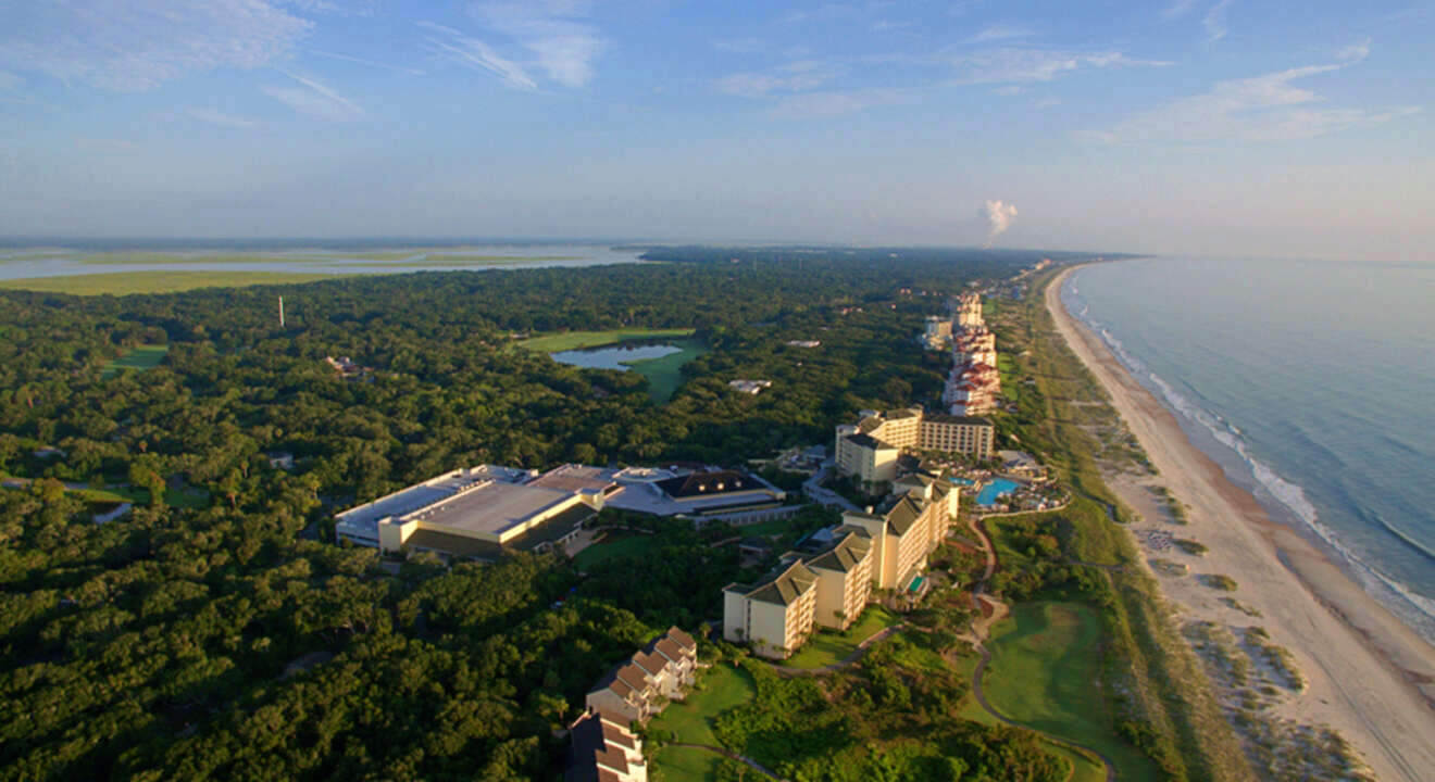 Aerial view of a coastal resort with multiple buildings, surrounded by trees and bordering a sandy beach with the ocean on the right.