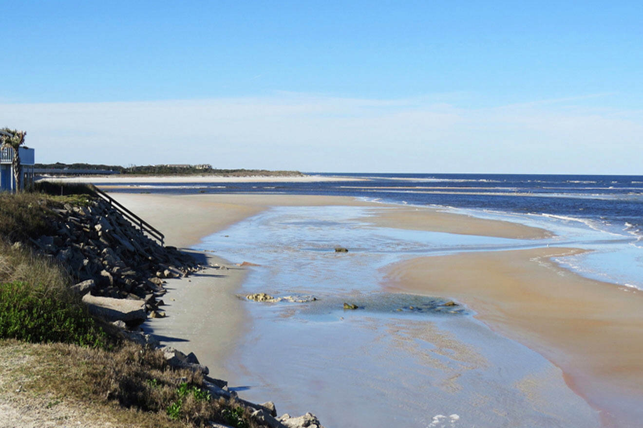 A serene beach with a flat shoreline, shallow water spanning across the sand, and a wooden staircase leading to the beach. The sky is clear and blue.