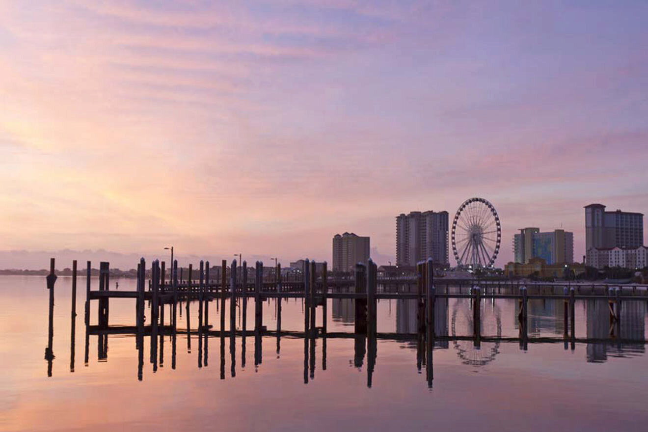 Sunset view of a calm waterfront with a pier, city skyline, and Ferris wheel in the background.