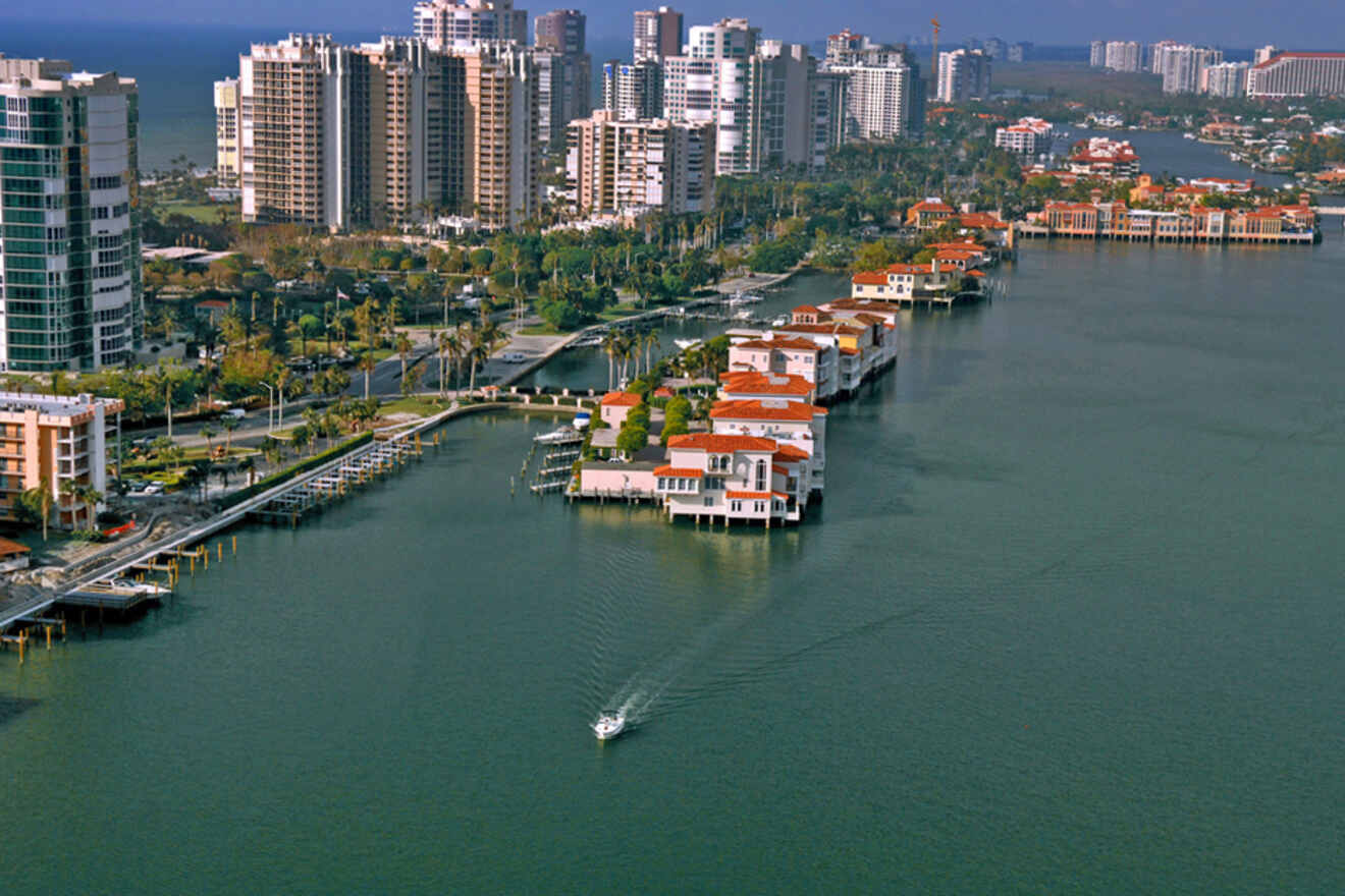 A small boat navigates a waterway lined with high-rise buildings and luxury homes, under a clear sky in Naples