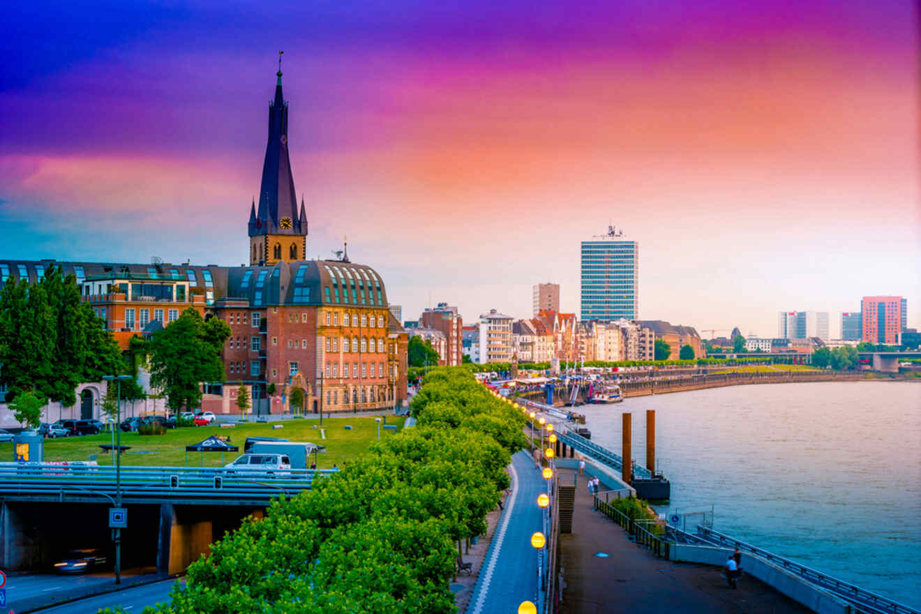 Cityscape with a church tower, historic buildings, and a river promenade at sunset.