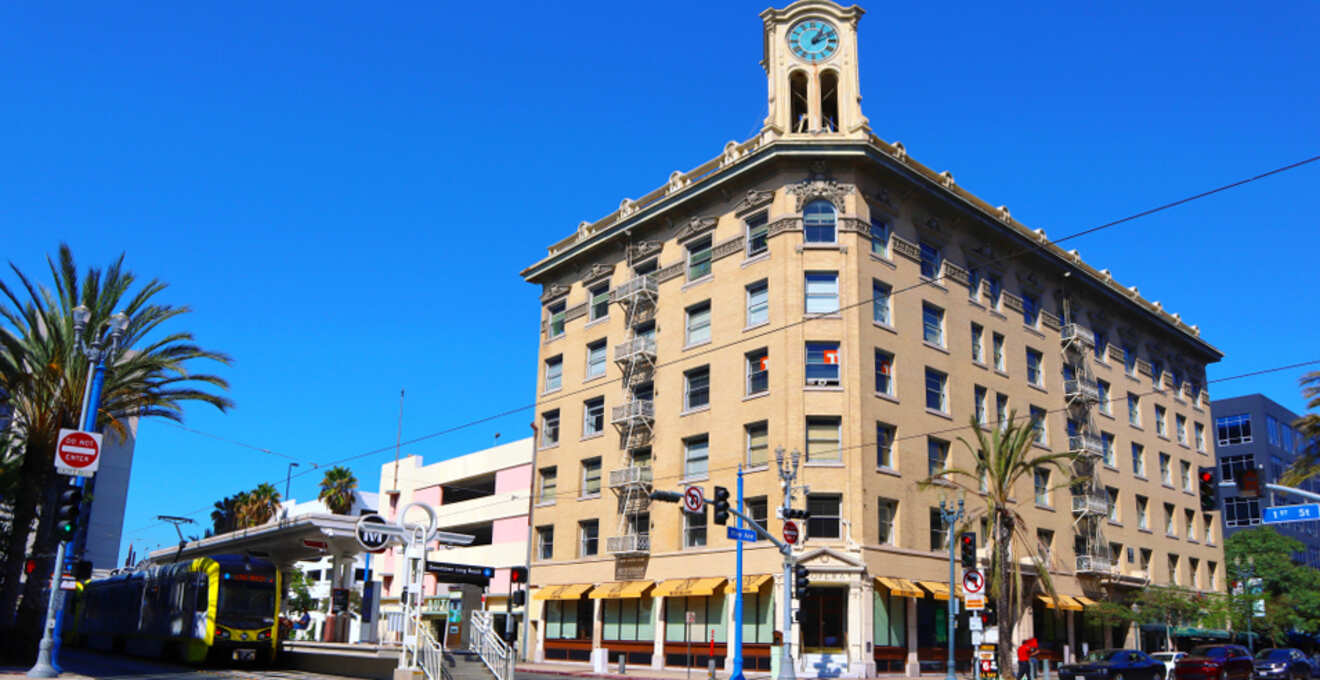 Historic clock tower building at the corner of a sunny downtown street in East Village Arts District, Long Beach, with a modern tram passing by and palm trees against a clear blue sky.