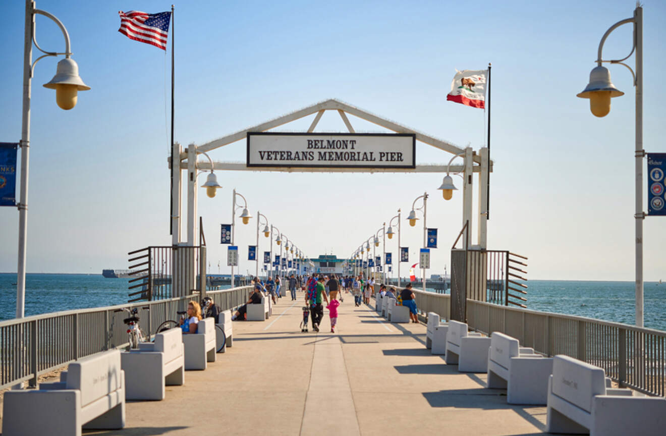 Visitors stroll down Belmont Veterans Memorial Pier in Long Beach, flanked by American and Californian flags, with the ocean extending into the horizon under a clear blue sky