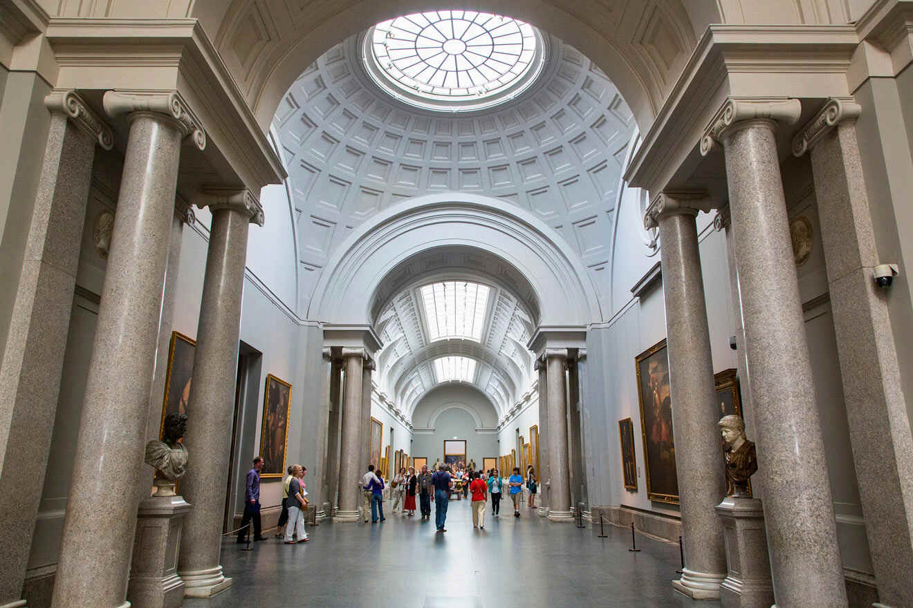 Interior of a grand museum gallery in Prado museum with visitors admiring artworks, featuring classical architecture with columns and a vaulted glass ceiling.