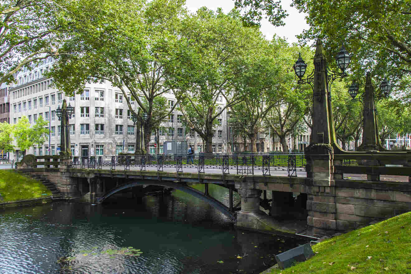 Bridge over a tree-lined canal in an urban setting, with historic buildings in the background.