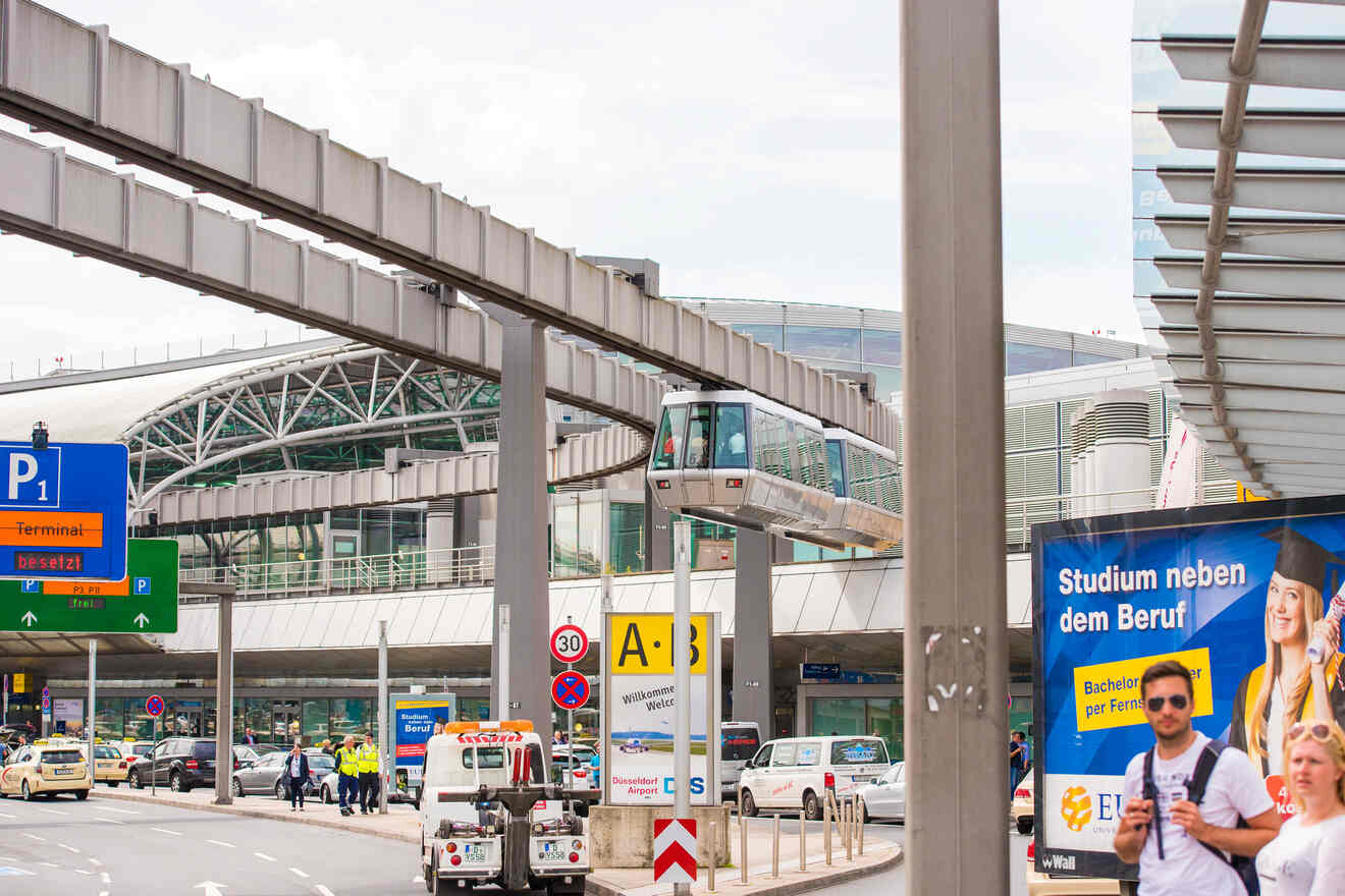 Airport scene with elevated train, parked cars, and people walking. Signs include directions and an advertisement.