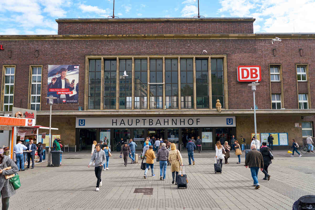 People walk in front of a train station labeled "Hauptbahnhof" with a large glass entrance, a DB sign, and nearby shops.