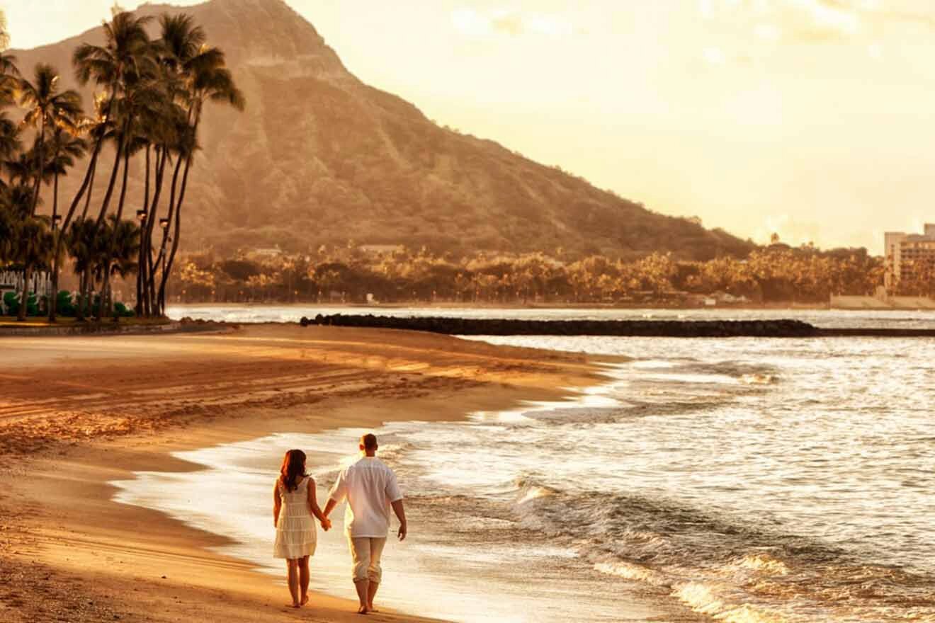 A couple holds hands while walking along a beach with palm trees and a mountain in the background during sunset.