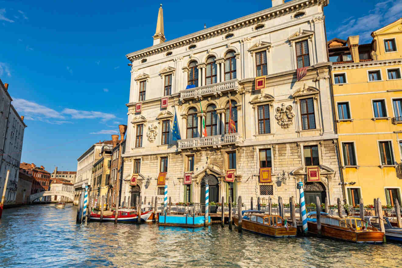 Historical building with arched windows, balconies, and flags, situated on a canal with docked boats in Venice, Italy under a clear blue sky.