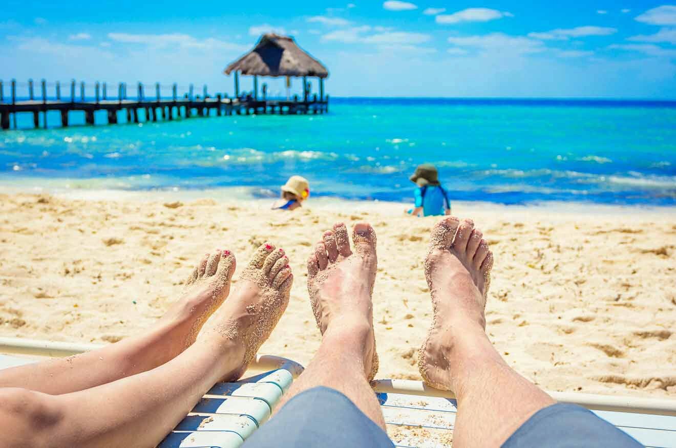 two people sitting on a beach with their feet in the sand and children in the background