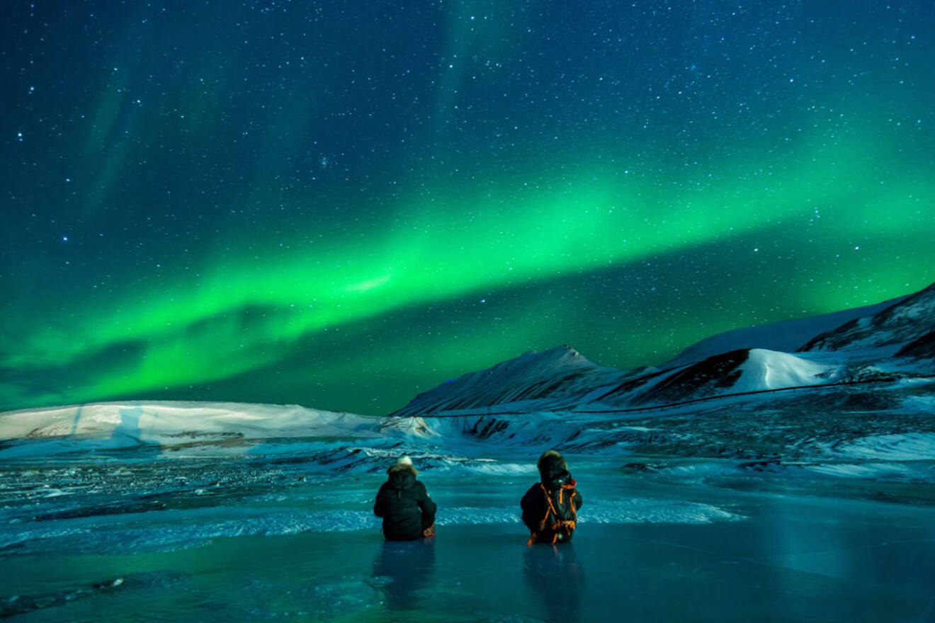 Two people sitting on ice, watching the vibrant green Northern Lights in a snowy, mountainous landscape.