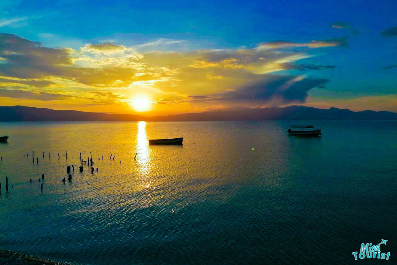 Sunset over a calm lake with two boats floating and wooden posts emerging from the water. A mix of blue, orange, and yellow hues fills the sky. "Miss Tourist" logo is in the bottom right corner.