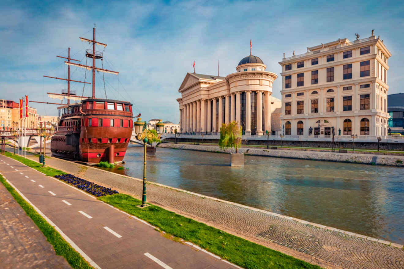 A red ship structure is on the riverside next to neoclassical buildings and a paved path under a blue sky.