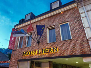 Brick building with signs reading "Hotel Bern," featuring flags and two windows, set against a blue sky background.