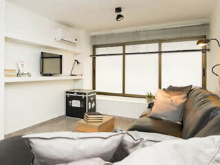 Modern living room with a black couch, pillows, a small wooden table, a TV mounted on a white wall, and a large window with blinds.