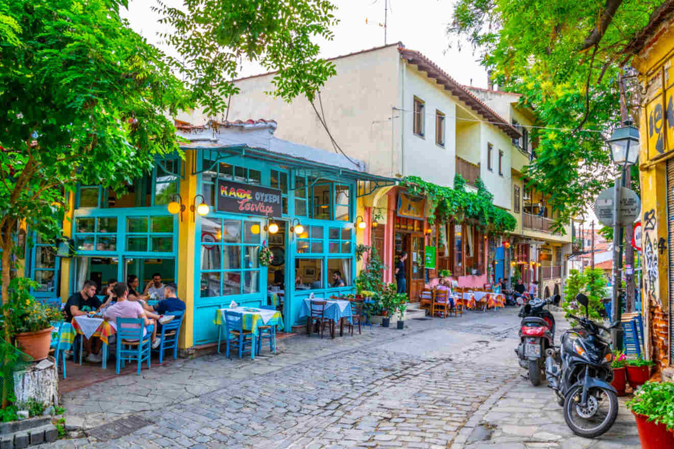 Colorful street scene with people dining outside a vibrant café, surrounded by greenery and quaint buildings. Motorcycles are parked along the cobblestone street.