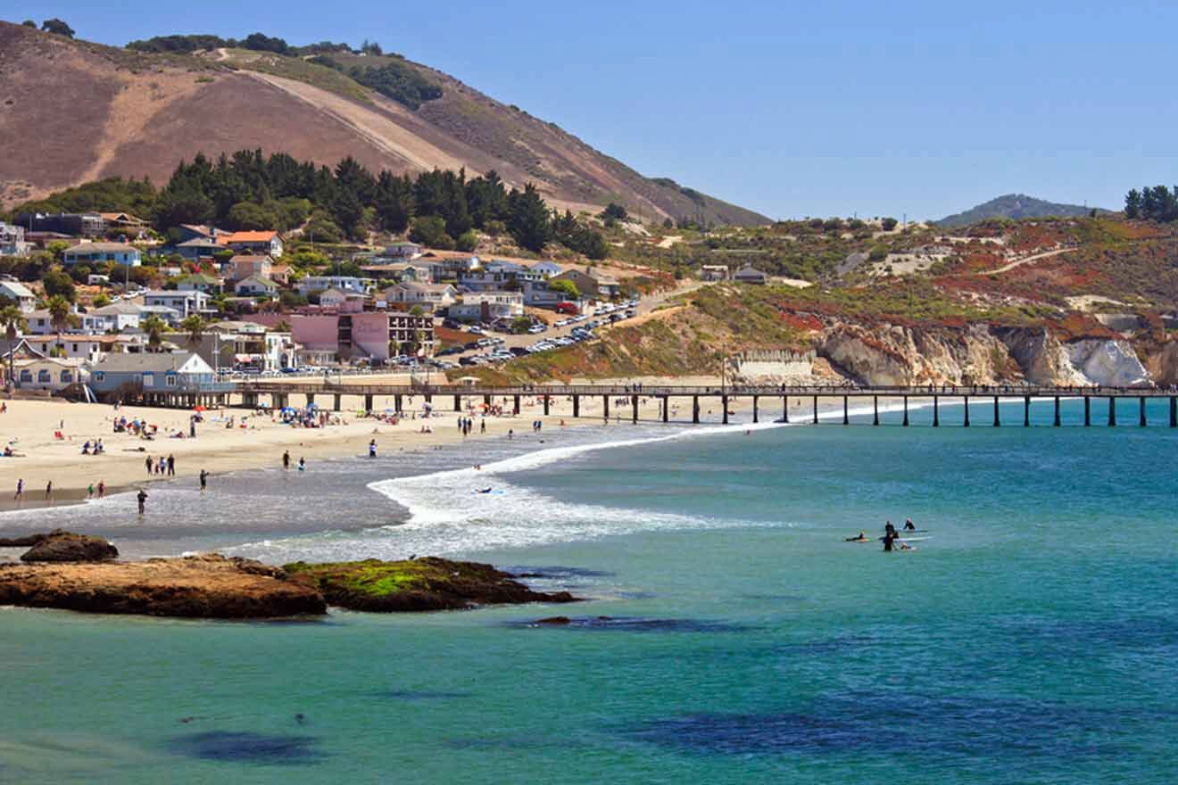 a group of people on a beach next to a pier with houses and hill in the background