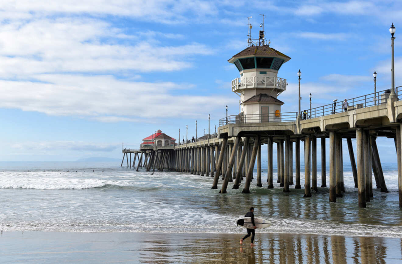 a person with surfboard walking on a beach next to a pier