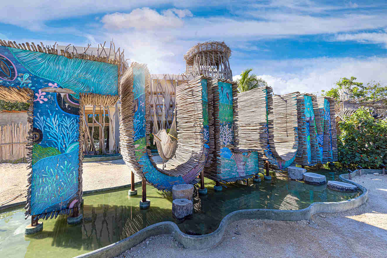 A large, colorful sign spelling "TULUM" made of wood and decorated with blue and green designs stands in front of a rustic building under a partly cloudy sky.