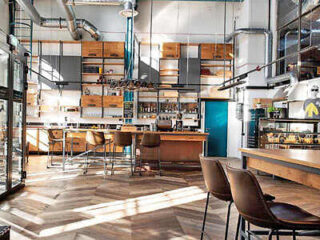 Modern café interior with high ceilings, large windows, wooden floors, and industrial design elements. There are wooden tables, chairs, and a bar area with shelves and various items.