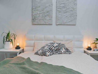 A neatly made bed with patterned pillows and a green blanket. Two abstract artworks hang above, flanked by potted plants and lit lamps on side tables.