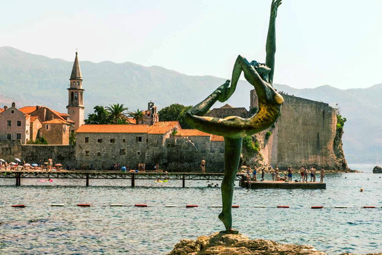 Statue of a dancer on a rock by the sea, with historic buildings and a church tower in the background.