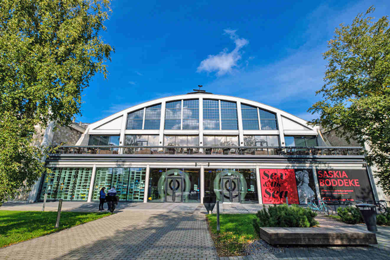 Modern glass building with an arched roof, trees on each side, and a banner on the right displaying text. Two people walk towards the entrance. Clear blue sky in the background.