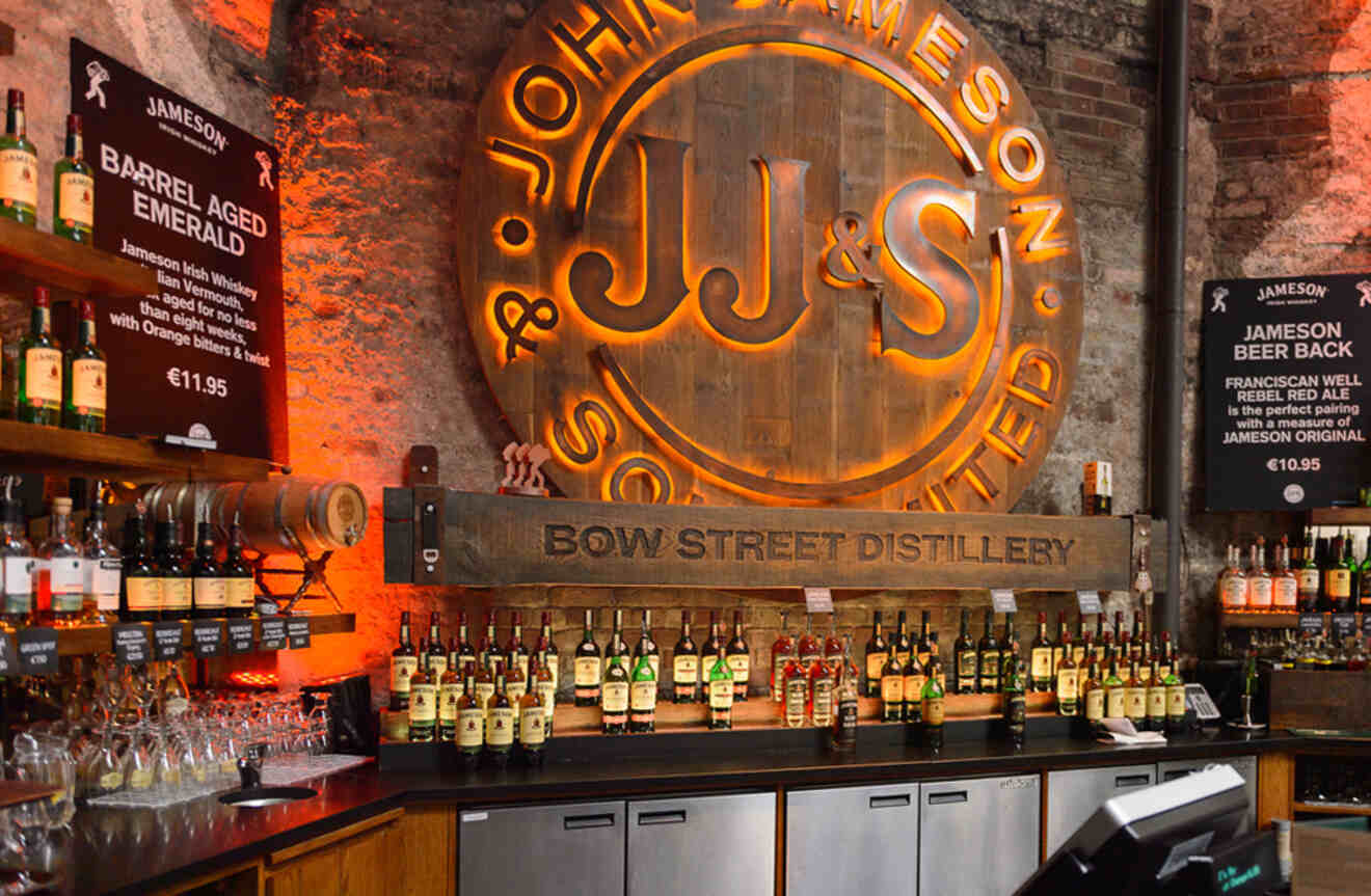 The interior of an Irish whiskey distillery bar with a large, illuminated sign of 'Jameson' and a selection of whiskey bottles on the counter.