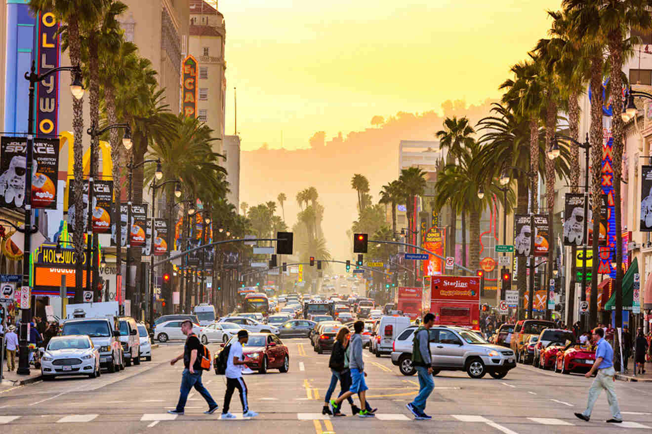 A vibrant street scene at sunset on Hollywood Boulevard with pedestrians crossing, neon signs, and palm trees against a hazy sky.
