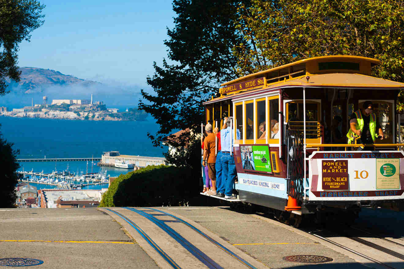 A historic San Francisco cable car filled with passengers, traveling down a steep street with Alcatraz Island and the Marina in the distant background under a clear blue sky.