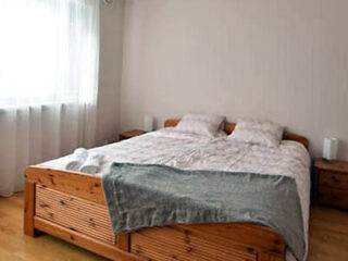 A neatly made wooden bed with two pillows and a folded blanket in a bright room with a window and two small bedside tables.