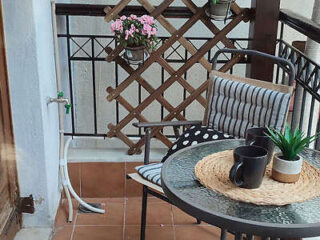 A small balcony with a round glass table, two black mugs, and a potted plant. A chair with striped cushions and a wooden lattice holding pink flowers are also visible.