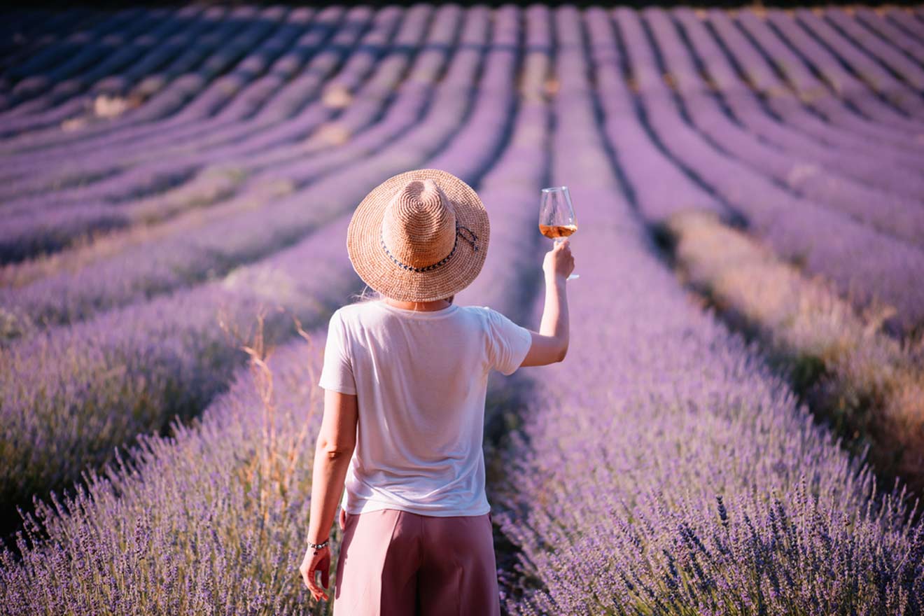 A person in a straw hat and white shirt holds a glass of wine while standing in a blooming lavender field.