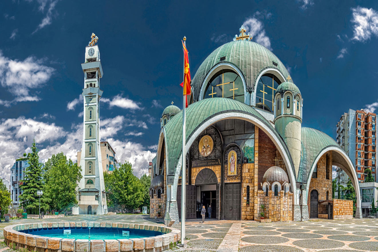 A domed church with arched entrances sits beside a clock tower. A national flag is in the foreground, and a round fountain is in the plaza.