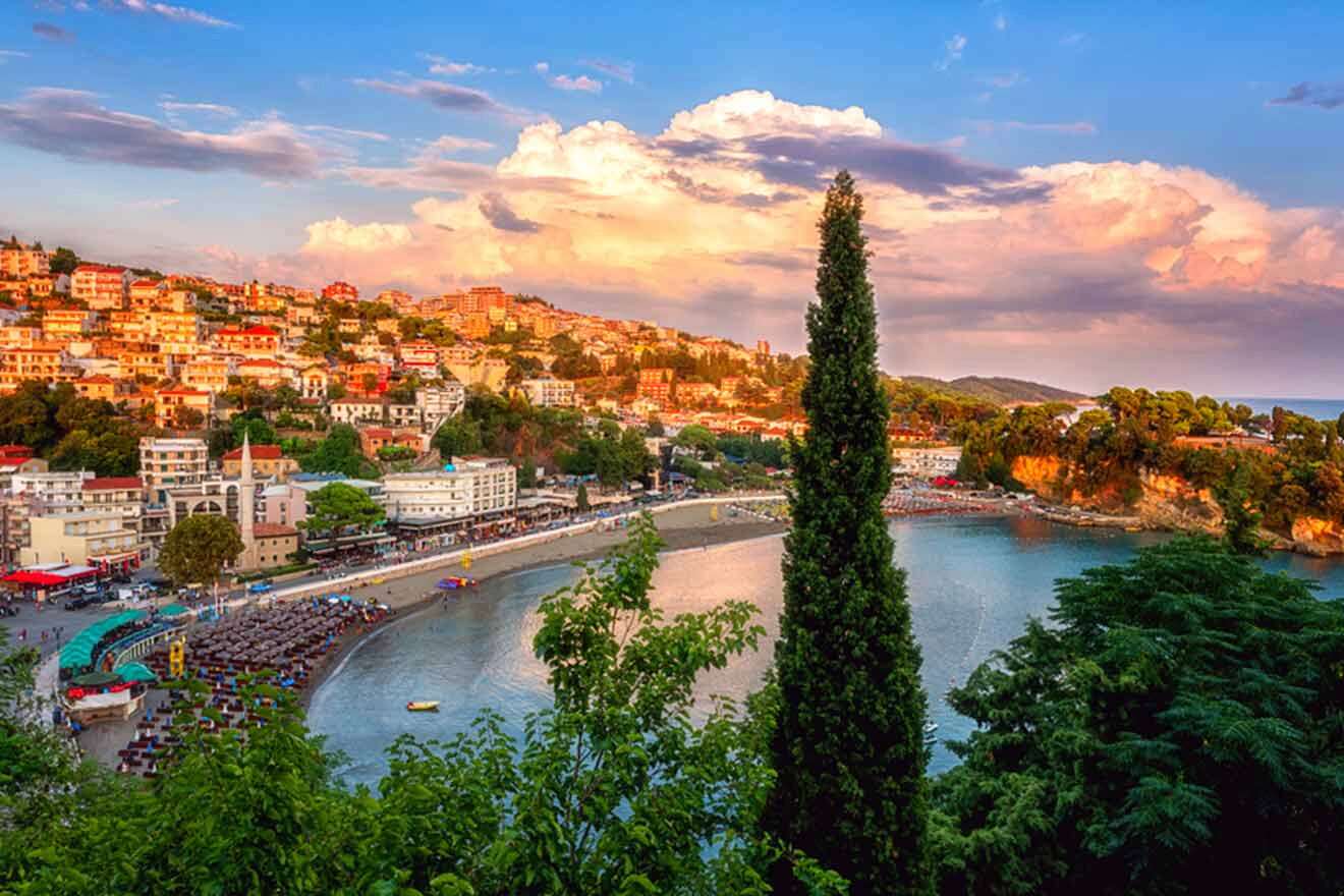 Elevated view of a coastal town with colorful buildings, a beach, and a calm bay. A tall cypress tree stands in the foreground, framed by a vibrant sky with clouds.