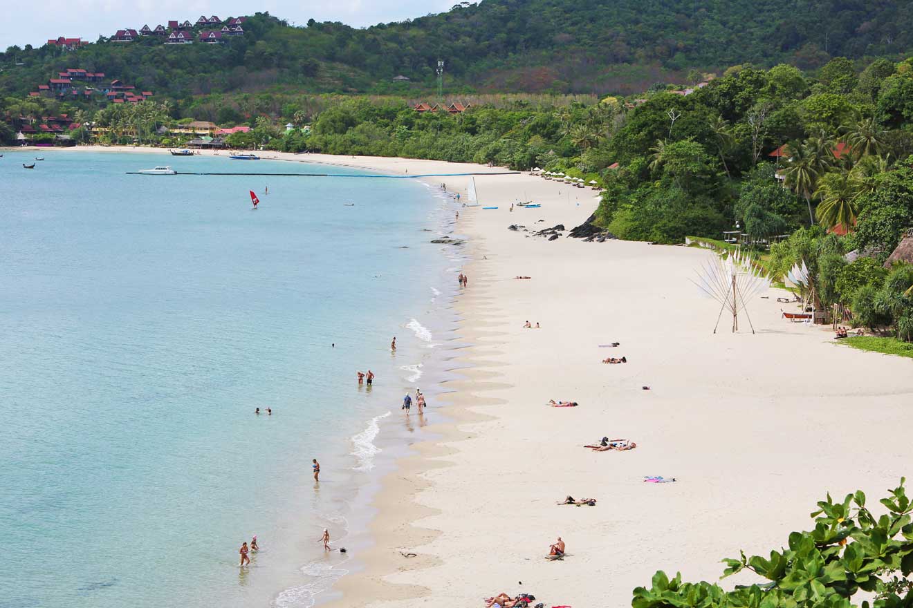 View of a wide sandy beach with visitors enjoying the sun, bordered by a calm sea on one side and lush green hills with scattered resorts on the other