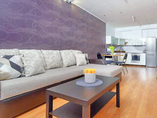 Modern living room with a gray sofa, patterned accent wall, and wooden floor. A black coffee table and a kitchen area with stainless steel appliances are visible in the background.