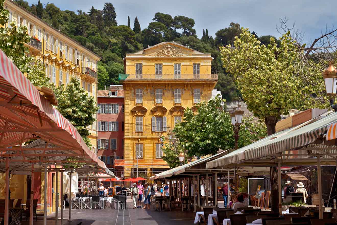 Busy outdoor market scene in Nice with shaded stalls, diners at outdoor cafes, and historic yellow buildings