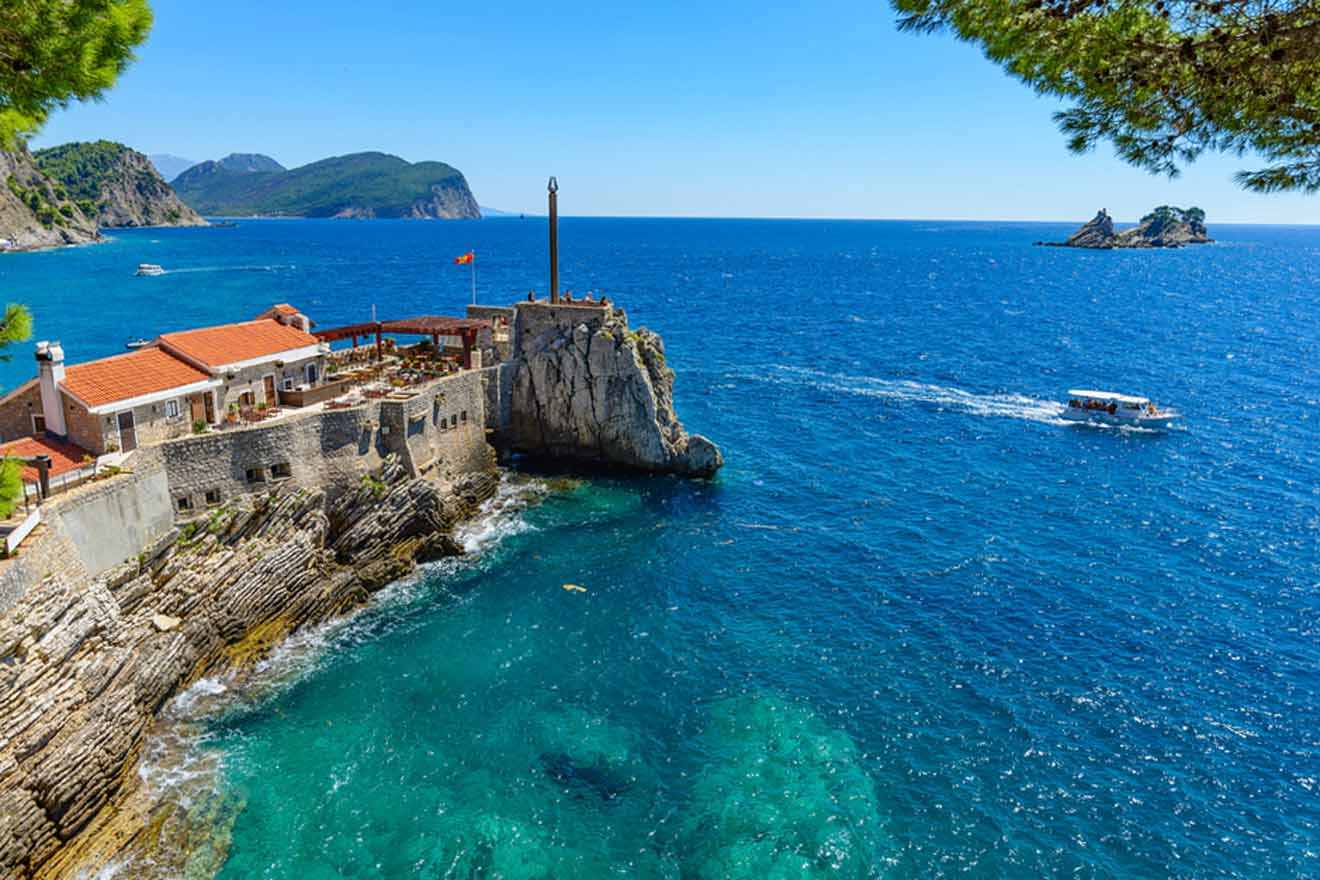 Coastal scene with a stone building on a cliff overlooking the sea, a small boat on the water, and distant rocky islets on a clear, sunny day.