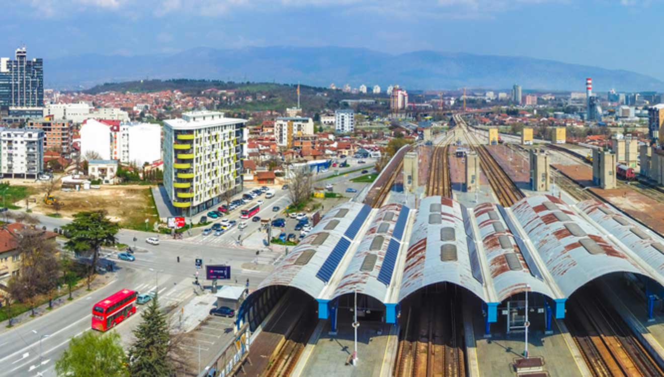 Aerial view of a train station with multiple tracks and platforms. Surrounding urban area includes buildings, a red bus, roads, and distant hills under a partly cloudy sky.