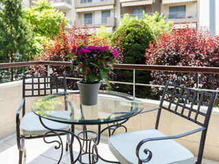 A glass table with a potted plant is flanked by two metal chairs on a balcony overlooking greenery and adjacent buildings.