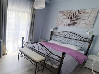 A bedroom with a wrought iron bed, pink and gray bedding, a bench at the foot, two nightstands, a leafy wall art, and a large window with sheer curtains.