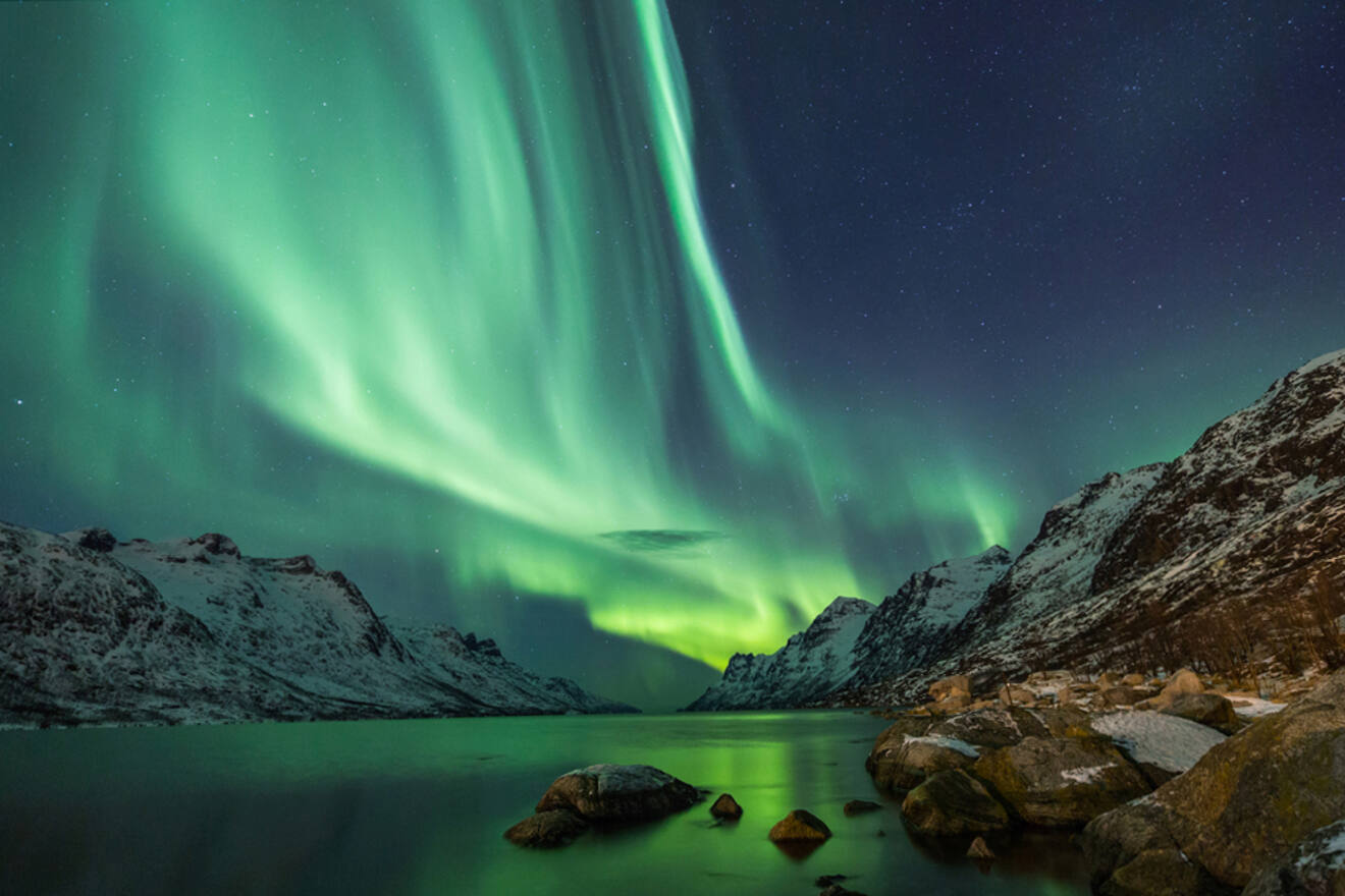 a body of water next to rocks and snowy field with aurora borealis in the sky