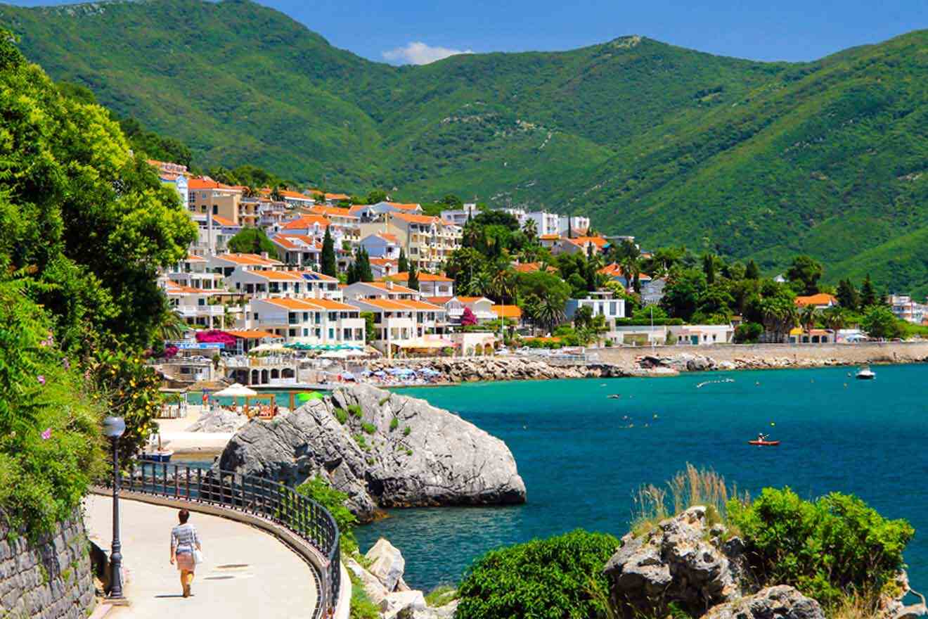 A scenic coastal village with red-roofed buildings nestled by green hills, overlooking a turquoise sea. A person walks along a path beside the water.