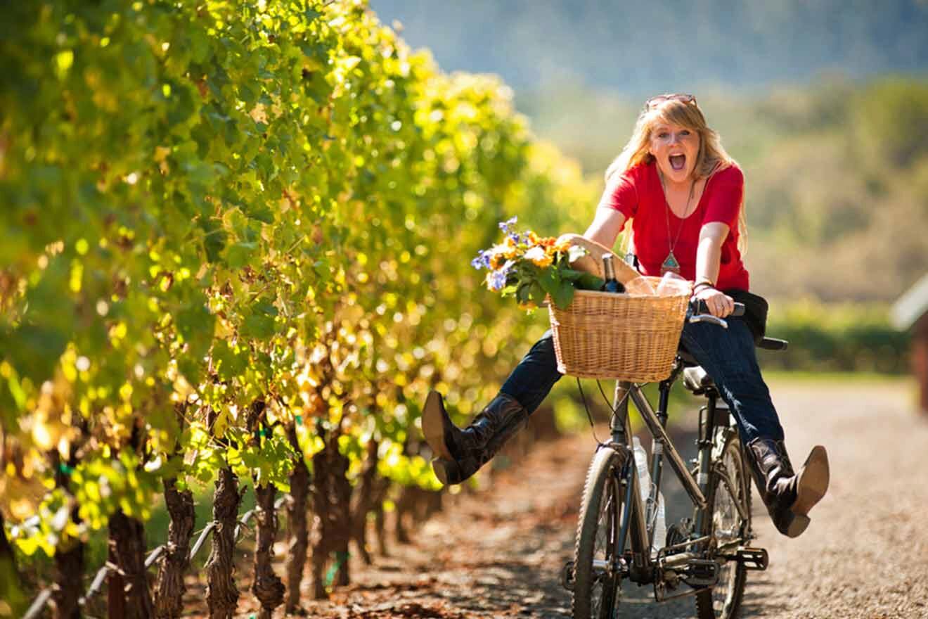 a woman riding a bike with a basket full of flowers and a bottle of wine