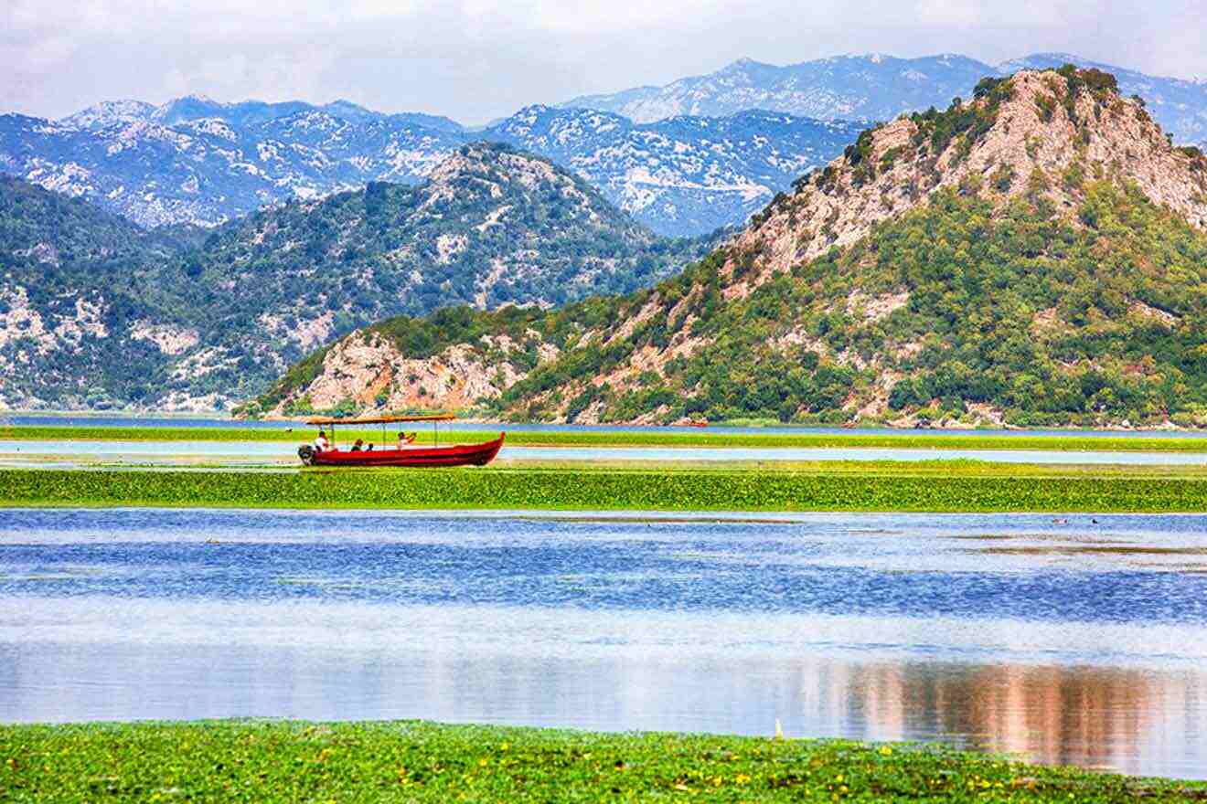 A red boat floats on a lake surrounded by lush green hills and distant mountains under a cloudy sky.