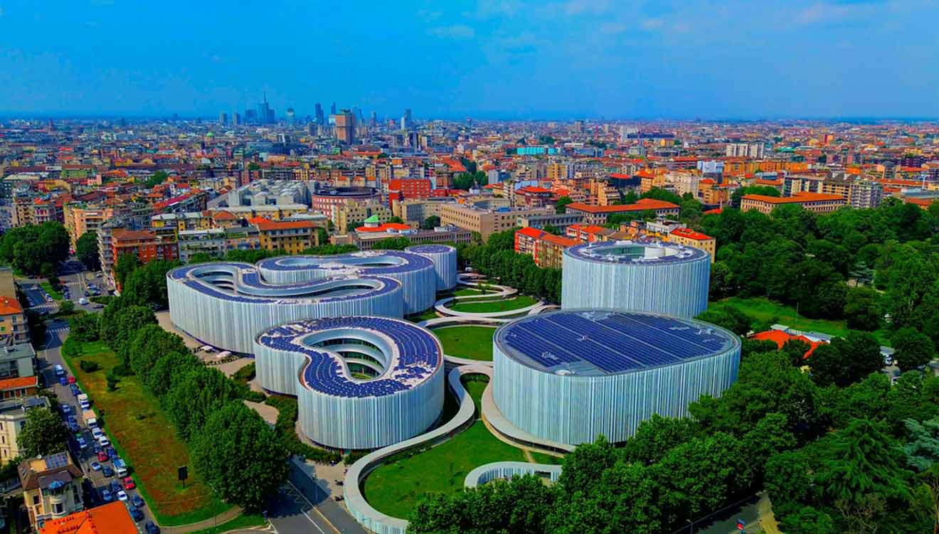 Aerial view of the modern, serpentine-shaped Milan Convention Centre (MiCo) surrounded by lush greenery, with the Milan skyline in the distance under a clear blue sky