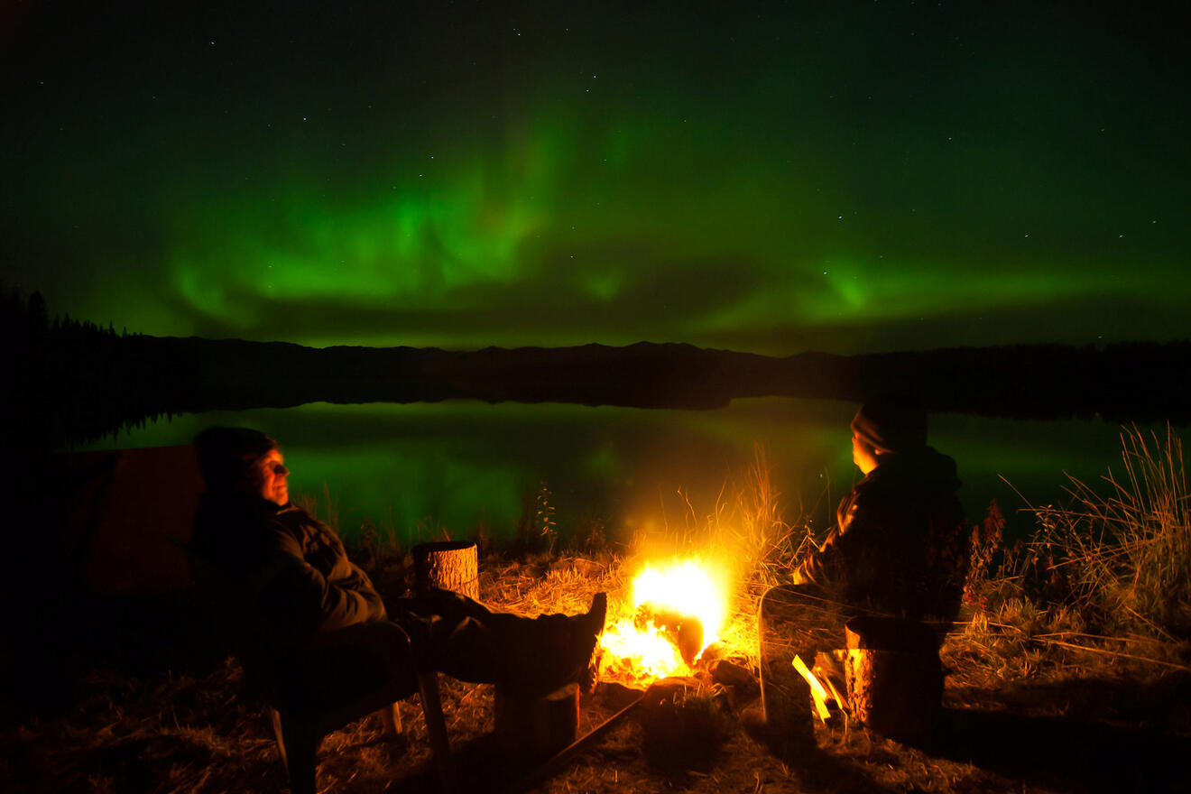 people getting warm by the fire next to a body of water watching the northern lights